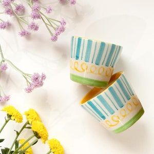 Pu&ograve; includere: Due piccole tazze in ceramica con motivi a strisce blu e bianche e accenti gialli. Le tazze sono su uno sfondo bianco con fiori gialli e viola.