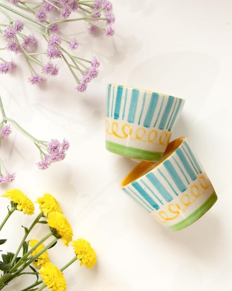 A vibrant set of 2 hand-painted ceramic shot glasses/espresso cups, each holding 75ml, with colorful designs, placed among fresh flowers, perfect for elevating any barware collection.