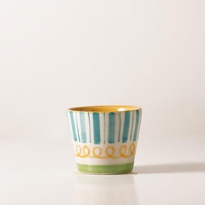 Pu&ograve; includere: Una piccola tazza in ceramica bianca con un bordo giallo e strisce blu e verdi. La tazza ha un design ondulato giallo intorno alla parte inferiore.