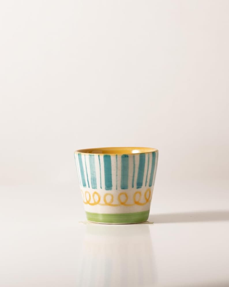 Pu&ograve; includere: Una piccola tazza in ceramica bianca con un bordo giallo e strisce blu e verdi. La tazza ha un design ondulato giallo intorno alla parte inferiore.