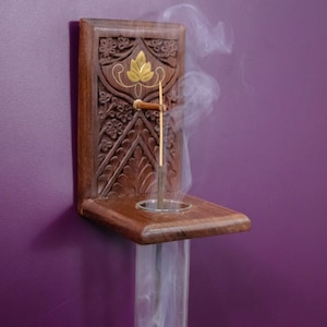 May include: A wooden incense holder with intricate carvings and a gold leaf design. A single incense stick is burning inside the holder, with smoke rising from the top.