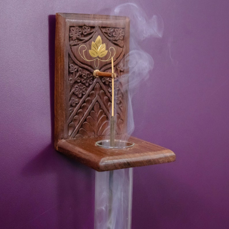 May include: A wooden incense holder with intricate carvings and a gold leaf design. A single incense stick is burning inside the holder, with smoke rising from the top.