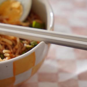 May include: A close-up of a bowl of noodles with a soft-boiled egg on top. The bowl is white with a checkered pattern in orange and white. A pair of chopsticks is resting on the rim of the bowl.