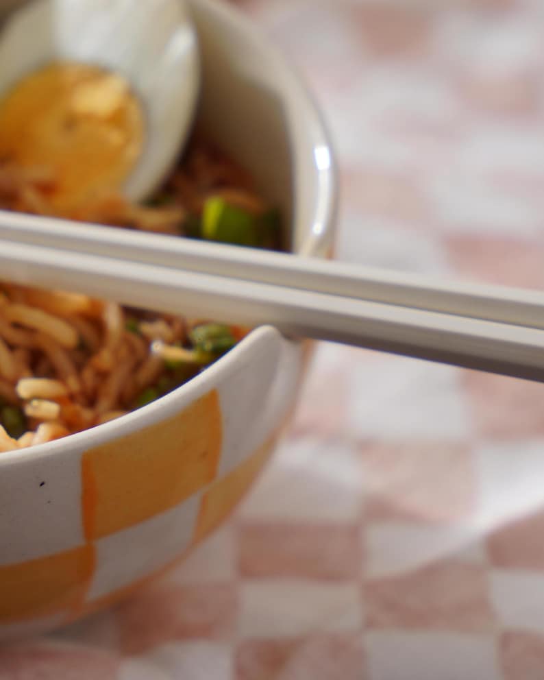 May include: A close-up of a bowl of noodles with a soft-boiled egg on top. The bowl is white with a checkered pattern in orange and white. A pair of chopsticks is resting on the rim of the bowl.