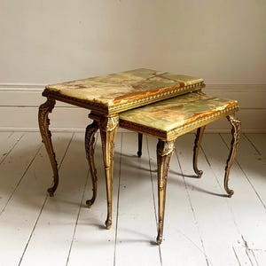 May include: A set of two nesting tables featuring rectangular tops made of polished green and brown onyx stone. The tables have ornate gold-coloured metal frames with curved, decorative legs and detailed trim along the edges of the stone tops.
