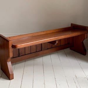 May include: A long, low-profile wooden bench featuring a warm brown finish, a flat rectangular seat, and a vertical slatted backrest. The bench is supported by two solid side panels with a notched cutout design at the base of each end.