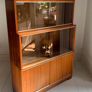 May include: A tall wooden cabinet featuring a warm brown finish with a visible wood grain. The unit consists of two upper sections with clear sliding glass doors and a lower section with solid wooden sliding doors, all framed in a light metal trim.