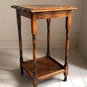 May include: An antique wooden side table with a square top and a lower shelf. The table has four turned legs and is made of a warm, medium-toned wood. The table is approximately 61 cm tall.