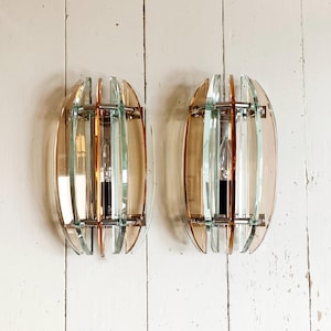May include: A pair of vintage wall sconces with an oval design. Each sconce features a clear glass bulb, surrounded by vertical glass panels in shades of amber and clear, held together by a silver metal frame. The sconces are mounted on a white wooden wall.