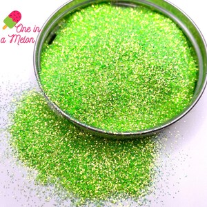 May include: A close-up of a green glitter with a fine shimmer. The glitter is in a small metal container and is spilling out onto a white surface.