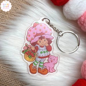 May include: A clear acrylic keychain featuring a cartoon character with a pink strawberry hat and dress, holding a basket of strawberries. A pink cat is beside the character. The keychain has a silver chain and ring. The text "Rocki Road Creations" is in the upper left corner.