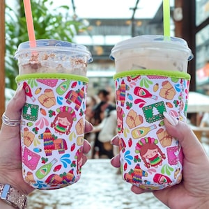 May include: Two clear plastic cups with iced drinks and straws are held in festive, patterned drink sleeves. The sleeves feature a white background with colourful illustrations of a girl, piñatas, and other celebratory icons. The sleeves have a green trim.
