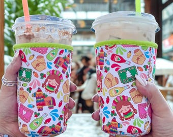 May include: Two clear plastic cups with iced drinks and straws are held in festive, patterned drink sleeves. The sleeves feature a white background with colorful illustrations of a girl, piñatas, and other celebratory icons. The sleeves have a green trim.