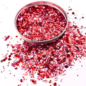May include: A small container filled with red and pink glitter. The glitter is scattered around the container on a white surface.