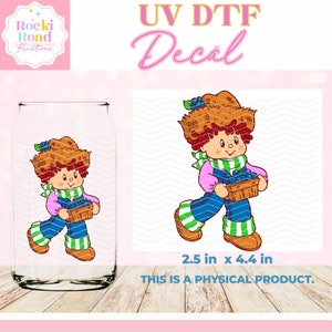May include: A UV DTF decal featuring a cartoon character with red hair, a brown straw hat, and a blue and green outfit. The character is holding a basket of blueberries. The decal is 2.5 inches by 4.4 inches.