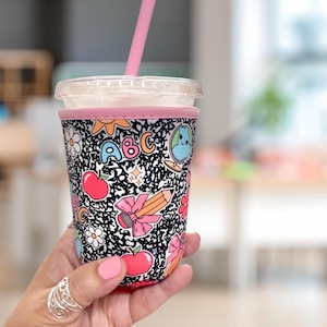 May include: A vibrant drink sleeve with a composition notebook pattern in black and white, adorned with school-themed illustrations. The sleeve, edged in pink, encases a clear plastic cup with a pink straw. The design incorporates apples, pencils, and the letters ABC.