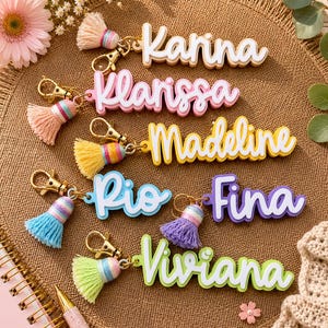 May include: A collection of six custom name keychains featuring layered acrylic lettering in white, pink, yellow, blue, purple, and green. Each keychain includes a gold-tone lobster clasp and a matching colored tassel with a striped accent band.