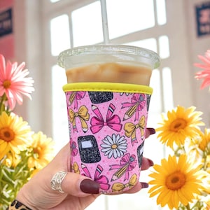 May include: A pink coffee cup sleeve with a yellow trim, decorated with bows, pencils, notebooks, and daisies. The sleeve holds a clear plastic cup with iced coffee and a straw. The background includes yellow and pink flowers.
