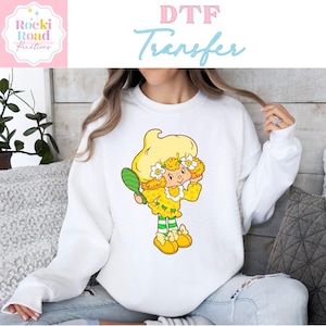 May include: White sweatshirt with a yellow and green cartoon character wearing a yellow dress and a flower crown. The character is holding a mirror and smiling. The text "DTF Transfer" is at the top of the image.