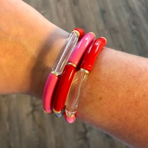 May include: A set of four bracelets in shades of pink, red, and clear. The bracelets have a rounded, tubular design with gold-colored accents. The clear bracelet has a vertical red line.