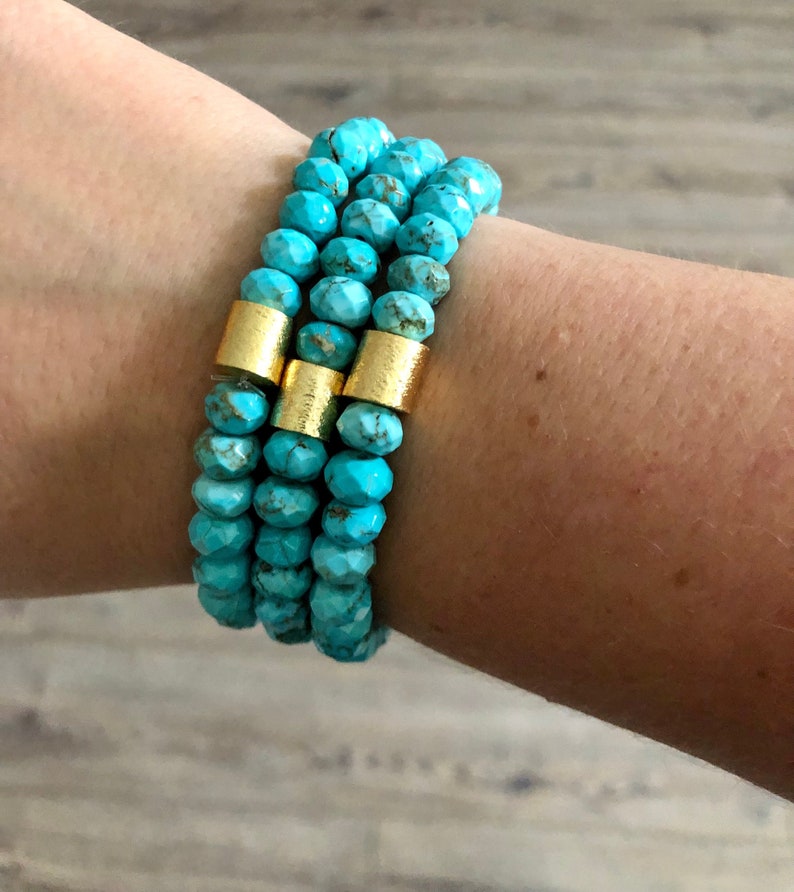 May include: Three turquoise beaded bracelets with gold spacers. The bracelets are stacked on a person's wrist.