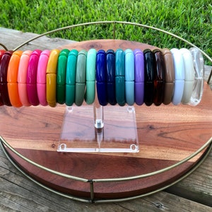 May include: A display of colourful bangle bracelets on a wooden tray with a gold metal rim. The bracelets are arranged in a circle on a clear acrylic stand. The bracelets are made from a variety of materials, including plastic, wood, and metal.