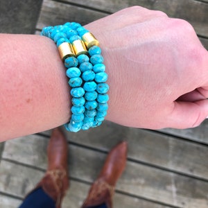 May include: A turquoise beaded bracelet with gold accents. The bracelet is layered and worn on a person's wrist.