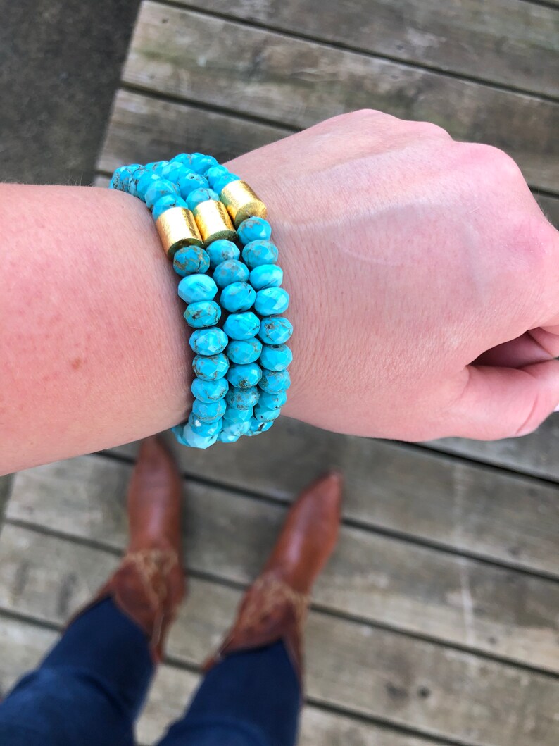 May include: A turquoise beaded bracelet with gold accents. The bracelet is layered and worn on a person's wrist.