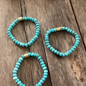 May include: Three turquoise beaded bracelets with gold accents. The bracelets are on a wooden surface.