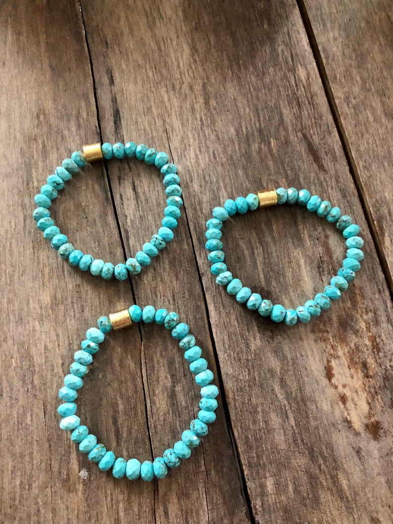 May include: Three turquoise beaded bracelets with gold accents. The bracelets are on a wooden surface.