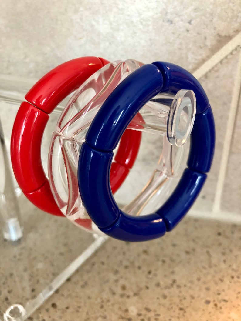 May include: Three bracelets, one red, one blue, and one clear, are stacked on top of each other. The bracelets are made of plastic and have a rounded shape.