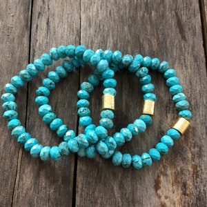 May include: Three turquoise beaded bracelets with gold accents. The bracelets are stacked on top of each other on a wooden surface.