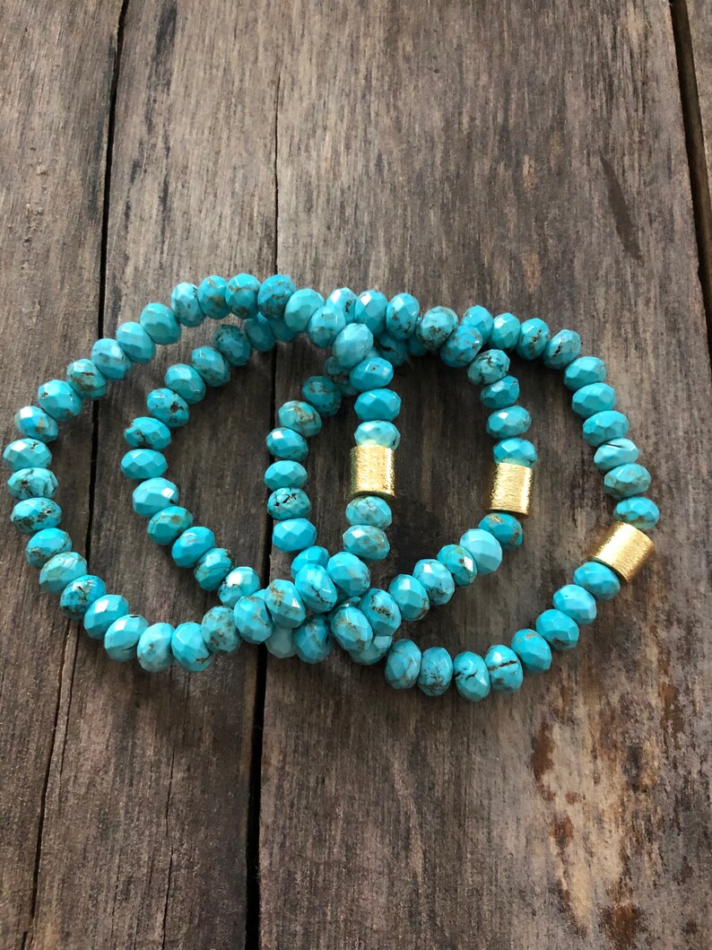 May include: Three turquoise beaded bracelets with gold accents. The bracelets are stacked on top of each other on a wooden surface.