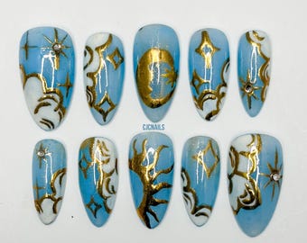May include: A set of ten almond-shaped press-on nails featuring a light blue base with white cloud-like accents and metallic gold celestial designs including moons, suns, and stars. Small clear rhinestones are embedded on select nails.