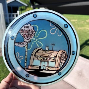 May include: A round, translucent disc featuring a graphic of The Krusty Krab restaurant from SpongeBob SquarePants. The design is framed by a black porthole border with blue rivets, set against a light blue background with white flower shapes.
