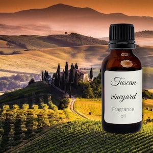 Puede incluir: Una botella de vidrio marrón con una tapa negra y una etiqueta blanca que dice "Tuscan vineyard Fragrance oil". La botella está delante de un paisaje escénico de colinas y viñedos.