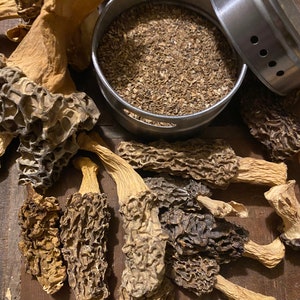 May include: A collection of dried morel mushrooms with a metal container filled with ground morels. The mushrooms vary in color from light tan to dark brown, with a distinctive honeycomb texture. The container has a lid with air holes.
