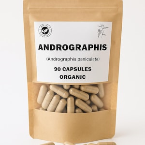 Puede incluir: Una bolsa de papel marrón con una ventana transparente que muestra cápsulas beige. La bolsa tiene una etiqueta blanca con el texto "ANDROGRAPHIS" y "90 CAPSULES ORGANIC". Algunas cápsulas están esparcidas frente a la bolsa.