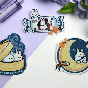 May include: Three illustrated stickers featuring cute white rabbits. The first sticker shows two rabbits in a steaming basket labelled "Steamed Buns". The second sticker shows a rabbit in a candy wrapper labelled "White Rabbit". The third sticker shows a rabbit fishing on a crescent moon with stars and a fish.