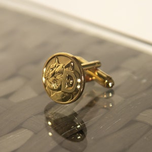 May include: Gold cufflinks with a cat face and the letter 'D' on the front.