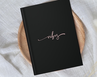 May include: A black notebook with the word "vibes" written in pink script on the front cover.