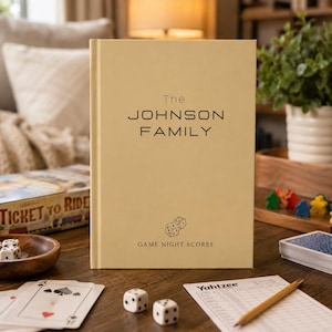 Puede incluir: Un libro de tapa dura de color beige con el texto The JOHNSON FAMILY impreso en negro, seguido de una pequeña ilustración lineal de dos dados y el texto GAME NIGHT SCORES. El libro está colocado en posición vertical sobre una superficie de madera.