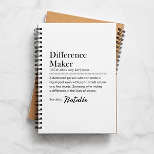 Puede incluir: Un cuaderno de espiral con una cubierta blanca que presenta texto negro que define el término Difference Maker como una persona dedicada que genera un gran impacto, seguido del texto See also: Natalie en una fuente cursiva negra.