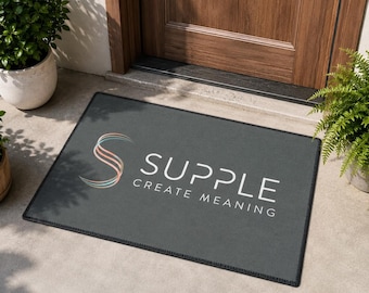 Puede incluir: Un felpudo rectangular de color gris oscuro que presenta un logotipo estilizado en forma de S con degradados en color melocotón y verde azulado a la izquierda, con el texto blanco "SUPPLE" en negrita y "CREATE MEANING" debajo.