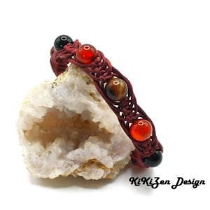 Op de afbeelding: Een rode, zwarte en bruine kralen armband met een geknoopt ontwerp. De armband ligt op een witte steen met een grotachtige opening.