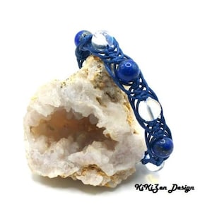 以下が含まれることがあります： ラピスラズリとクリアクォーツのビーズが付いた青と白のマクラメブレスレット。ブレスレットは、中央が空洞の白い岩の上に置かれています。