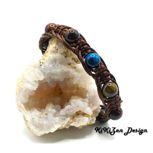 Op de afbeelding: Een bruin gevlochten armband met vijf edelsteen kralen. De kralen zijn rood, bruin, blauw, bruin en rood. De armband ligt op een witte steen met een holle kern.