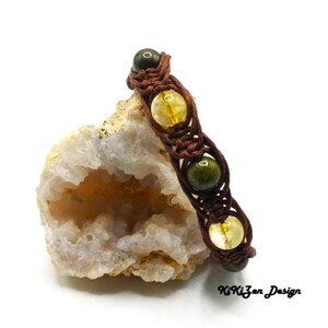 Pode incluir: Um bracelete de couro castanho com contas de pedras preciosas verdes e amarelas. O bracelete é trançado e tem um design simples. O bracelete é apresentado numa rocha branca.