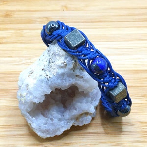 Pode incluir: Um bracelete tran&ccedil;ado azul com contas azul escuro, prateadas e pretas. O bracelete est&aacute; sobre uma rocha branca com um centro oco.