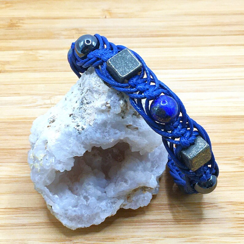 Pode incluir: Um bracelete tran&ccedil;ado azul com contas azul escuro, prateadas e pretas. O bracelete est&aacute; sobre uma rocha branca com um centro oco.